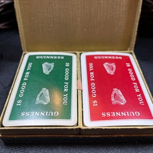 May include: A vintage Guinness playing card set in a gold-colored box. The cards are green and red, with the Guinness logo and the phrase "IS GOOD FOR YOU." Each card features a harp illustration.