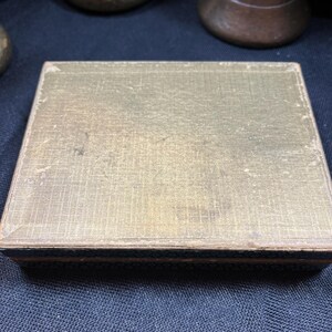 May include: A rectangular, antique box with a gold-colored, textured lid. The box has a dark blue border and appears to be made of a woven material. The box is sitting on a dark blue surface. Other antique objects are in the background.