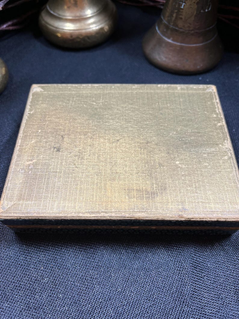May include: A rectangular, antique box with a gold-colored, textured lid. The box has a dark blue border and appears to be made of a woven material. The box is sitting on a dark blue surface. Other antique objects are in the background.