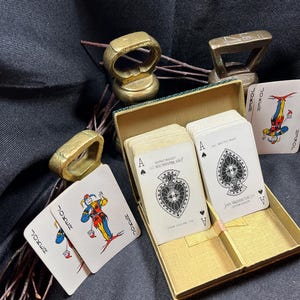 May include: A vintage playing card set with a gold-colored box, featuring cards with the Ace of Spades and Joker cards. The box is open, revealing the cards inside. Also, there are several gold-colored rings and a small gold-colored weight.