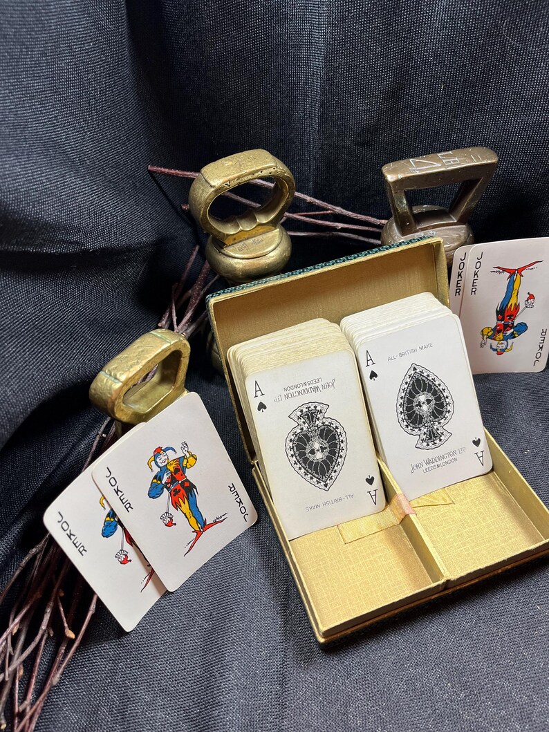 May include: A vintage playing card set with a gold-colored box, featuring cards with the Ace of Spades and Joker cards. The box is open, revealing the cards inside. Also, there are several gold-colored rings and a small gold-colored weight.
