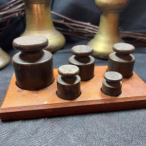 May include: A collection of antique brass weights of different sizes, displayed on a wooden tray. The weights are cylindrical with a rounded top. The tray is rectangular with a dark wood finish. Other brass objects are in the background.