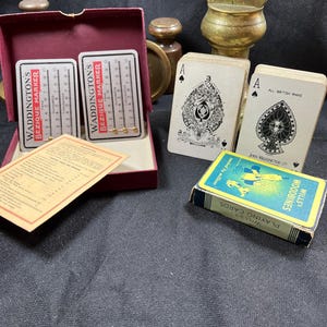 Könnte beinhalten: Eine Sammlung von Vintage-Spielkarten und Zubehör. Enthält zwei Waddington's Bezique Marker, ein Kartenspiel von Wills's Woodbines und zwei Pik-Ass-Karten. Die Artikel sind auf einer dunklen Oberfläche angeordnet.