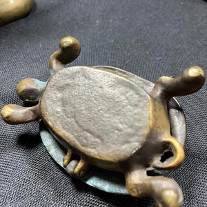 May include: A small, antique bronze turtle figurine with a weathered patina. The turtle has detailed legs, a textured shell, and a loop on the side. The bronze has areas of green oxidation, adding to its aged appearance.