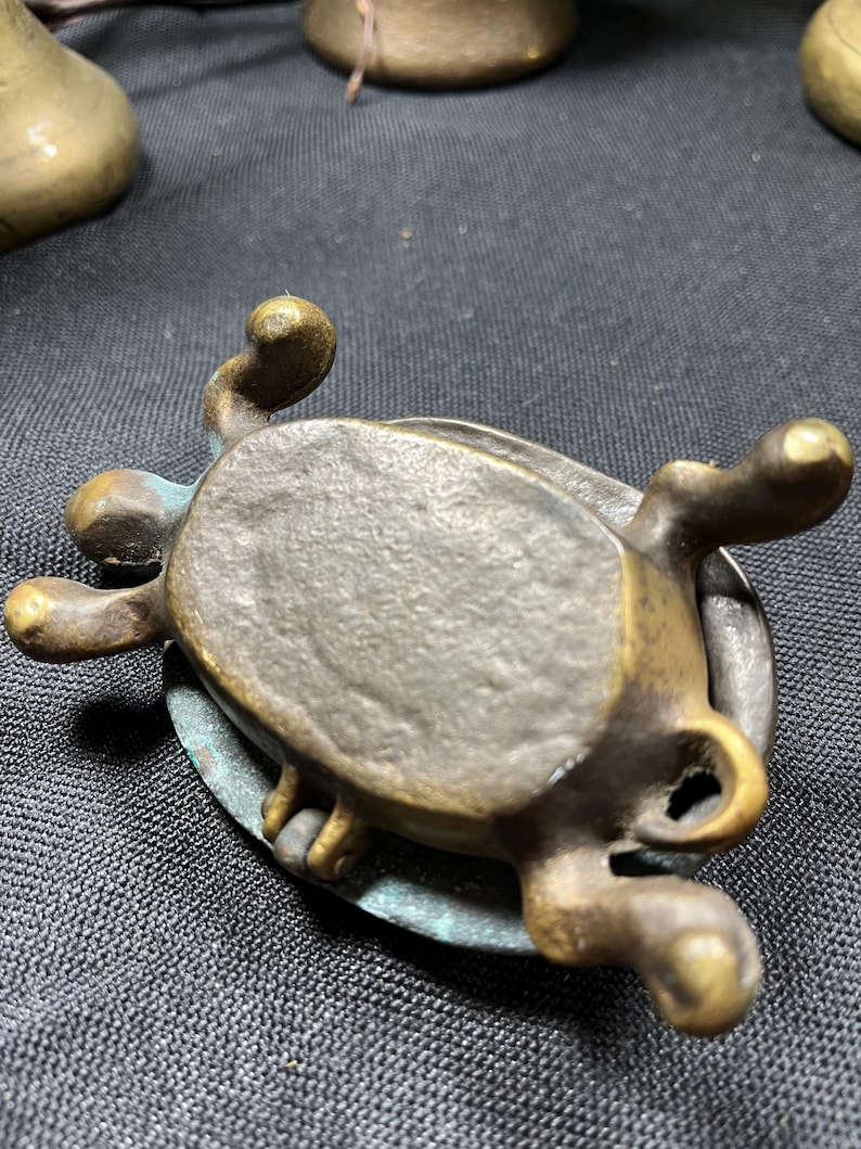 May include: A small, antique bronze turtle figurine with a weathered patina. The turtle has detailed legs, a textured shell, and a loop on the side. The bronze has areas of green oxidation, adding to its aged appearance.