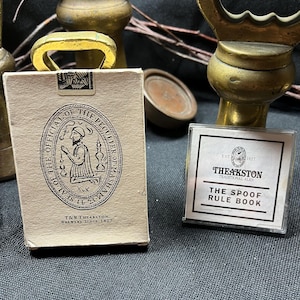 Könnte beinhalten: Eine Ansammlung von Messingobjekten und zwei kleine Schachteln. Eine Schachtel ist beige mit einem schwarzen Siegel und dem Text "T&R Theakston Brewers Since 1827". Die andere Schachtel ist transparent mit dem Text "The Spoof Rule Book" und "Theakston Traditional Ales."