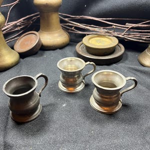 May include: Three small metallic mugs with single handles, featuring a rounded body and a flared rim. These miniature vessels have a smooth, muted finish and are arranged in a cluster, showcasing a consistent design with a circular base.