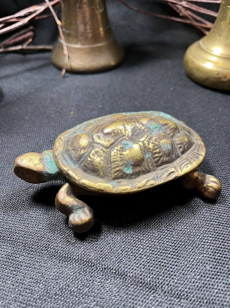 May include: A small, antique brass turtle figurine with a textured shell and detailed legs. The turtle has a weathered appearance with hints of green patina, suggesting age. The figurine is a decorative item.