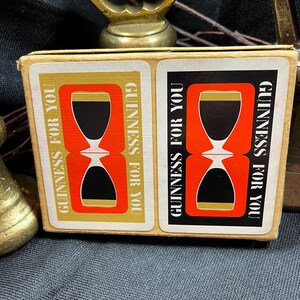 May include: A vintage Guinness For You matchbox featuring a red and black hourglass design with gold accents. The text "GUINNESS FOR YOU" is printed vertically on the sides. A brass bottle opener is visible in the background.