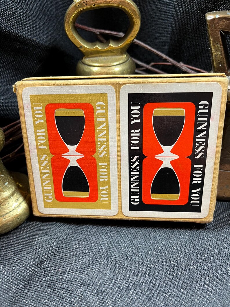 May include: A vintage Guinness For You matchbox featuring a red and black hourglass design with gold accents. The text "GUINNESS FOR YOU" is printed vertically on the sides. A brass bottle opener is visible in the background.