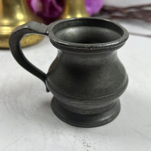 Puede incluir: Una peque&ntilde;a jarra de peltre antigua con asa. La jarra tiene un cuerpo redondeado y un borde acampanado. El asa es curva y est&aacute; unida al lateral. La jarra es de color gris oscuro y parece envejecida.