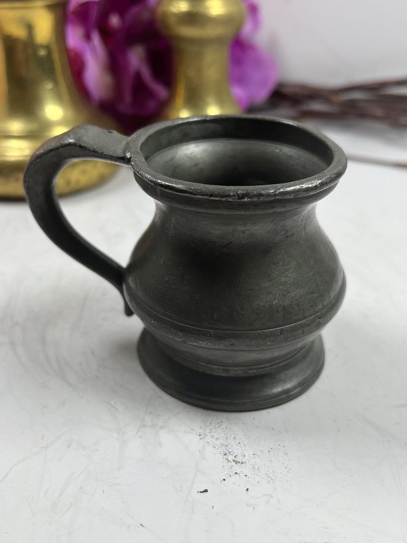 Puede incluir: Una peque&ntilde;a jarra de peltre antigua con asa. La jarra tiene un cuerpo redondeado y un borde acampanado. El asa es curva y est&aacute; unida al lateral. La jarra es de color gris oscuro y parece envejecida.