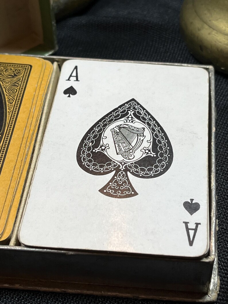 May include: Close-up of an Ace of Spades playing card. The card features a detailed black spade design with an ornate harp illustration in the center. The card is white with black accents and is part of a deck of cards.