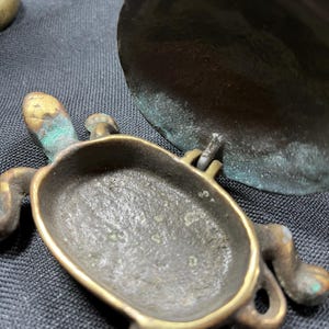 May include: A small, antique brass turtle-shaped container with a hinged lid. The turtle has detailed legs, head, and tail. The brass has a weathered appearance with hints of green patina, suggesting age and use. The container is open, revealing its interior.