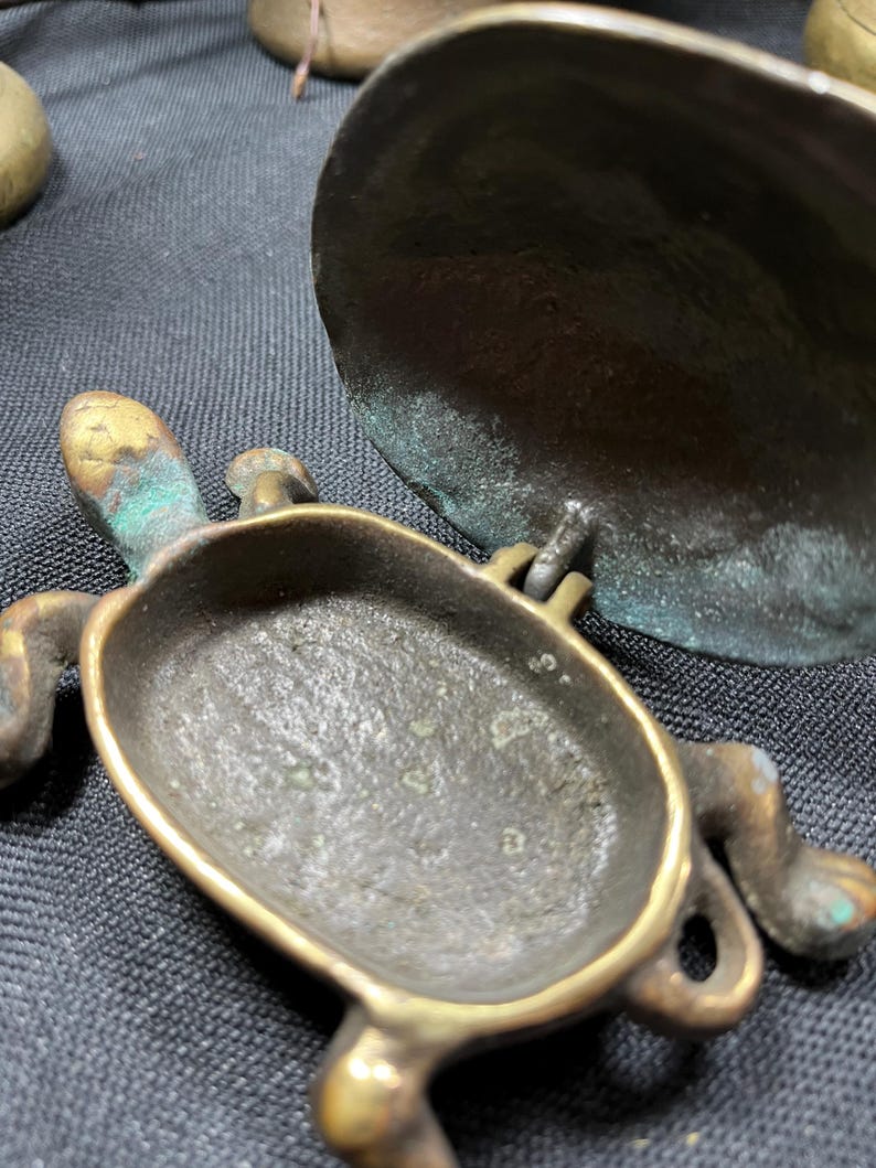 May include: A small, antique brass turtle-shaped container with a hinged lid. The turtle has detailed legs, head, and tail. The brass has a weathered appearance with hints of green patina, suggesting age and use. The container is open, revealing its interior.
