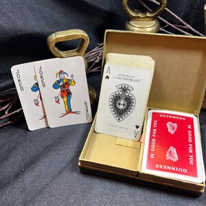May include: A deck of playing cards with a gold-colored box. Two joker cards are visible, along with a card featuring the Ace of Spades. A red card with the Guinness logo is also present. The cards are set against a dark background.