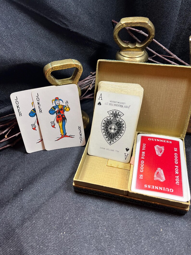 May include: A deck of playing cards with a gold-colored box. Two joker cards are visible, along with a card featuring the Ace of Spades. A red card with the Guinness logo is also present. The cards are set against a dark background.