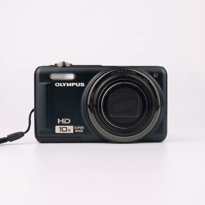 Peut inclure: Un appareil photo numérique Olympus noir avec un objectif argenté et une dragonne noire. L'appareil photo a un bouton argenté sur le dessus et le texte "Olympus" et "HD 10x Super Wide" imprimé sur le devant.