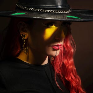 May include: A person with red hair wearing a black wide-brimmed hat with a silver chain and green accents. The person has yellow eyeshadow and red lipstick.