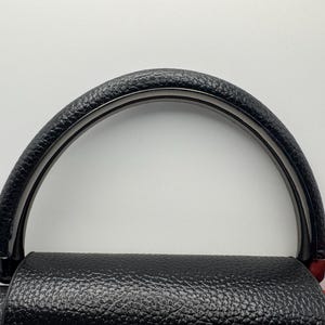 Pode incluir: Al&ccedil;a de bolsa preta com punho de couro texturizado e arco preto brilhante. A al&ccedil;a &eacute; presa a uma bolsa de couro preta com um detalhe vermelho. O design &eacute; simples e elegante.