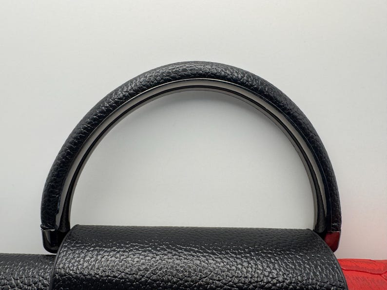 Pode incluir: Al&ccedil;a de bolsa preta com punho de couro texturizado e arco preto brilhante. A al&ccedil;a &eacute; presa a uma bolsa de couro preta com um detalhe vermelho. O design &eacute; simples e elegante.