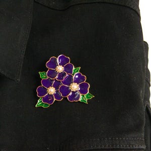 May include: A purple flower brooch with gold trim and pearl-like centers is pinned to a black denim jacket. The brooch features three flowers with green leaves.