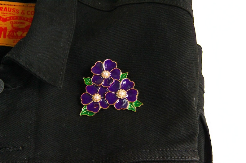 May include: A purple flower brooch with gold trim and pearl-like centers is pinned to a black denim jacket. The brooch features three flowers with green leaves.