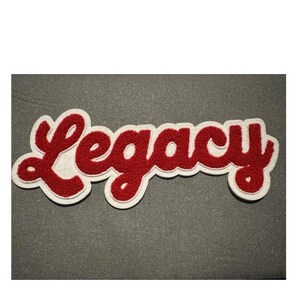 May include: A red and white embroidered patch with the word "Legacy" in a script font. The patch has a white border and is set against a gray background. The letters are in a bold, cursive style.