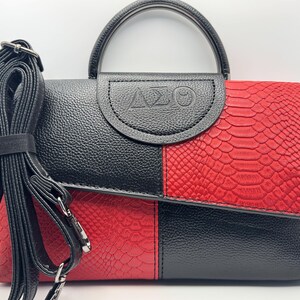 Pode incluir: Bolsa de m&atilde;o preta e vermelha com efeito pele de cobra. Possui uma al&ccedil;a superior e uma al&ccedil;a de ombro remov&iacute;vel. A aba &eacute; estampada com as letras &Delta;&Sigma;&Theta;. A bolsa &eacute; dividida em duas se&ccedil;&otilde;es com blocos de cores.