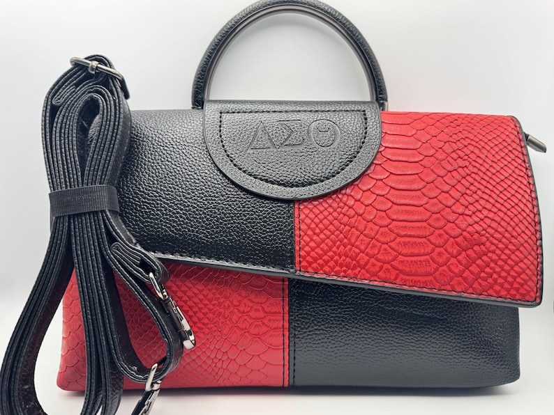 Pode incluir: Bolsa de m&atilde;o preta e vermelha com efeito pele de cobra. Possui uma al&ccedil;a superior e uma al&ccedil;a de ombro remov&iacute;vel. A aba &eacute; estampada com as letras &Delta;&Sigma;&Theta;. A bolsa &eacute; dividida em duas se&ccedil;&otilde;es com blocos de cores.