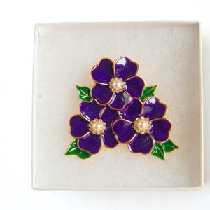 May include: A purple floral brooch with gold trim and pearl-like centers, presented in a white box. The brooch features three flowers with green leaves, creating a vibrant and elegant design. A perfect accessory for any outfit.