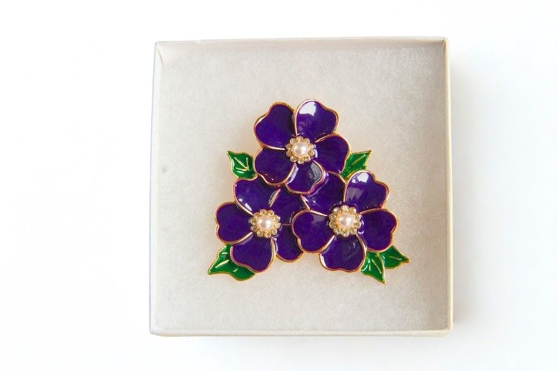 May include: A purple floral brooch with gold trim and pearl-like centers, presented in a white box. The brooch features three flowers with green leaves, creating a vibrant and elegant design. A perfect accessory for any outfit.