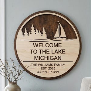 May include: A circular wooden sign with a dark brown frame and a light wood centre. The sign says "WELCOME TO THE LAKE MICHIGAN" with "THE WILLIAMS FAMILY EST. 2025 43.5°N, 87.3°W" below. A sailboat and trees are carved into the top.