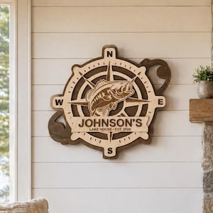 May include: Wooden compass-themed wall art with a fish illustration and the text "JOHNSON'S LAKE HOUSE EST. 2020". The artwork is mounted on a white shiplap wall. A sofa with fish-themed cushions and a mug that says "LIFE IS BETTER AT THE LAKE" are in the foreground.