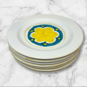 May include: A stack of white ceramic plates featuring a hand-painted design in the center. The pattern consists of a bright yellow flower surrounded by a teal blue circular border with small yellow dots, all set against a white background.