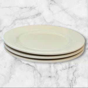 May include: A stack of three round, off-white ceramic dinner plates. Each plate features a smooth, glossy finish with a wide, flat rim and a slightly recessed center. The plates are uniform in color and shape, stacked neatly on top of one another.
