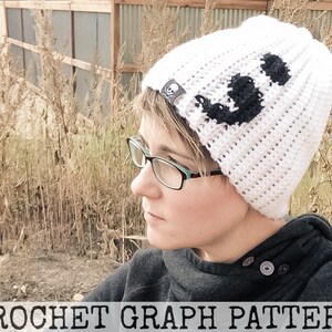 May include: White crochet beanie with a black skull and crossbones graphic. The beanie is being worn by a person with short brown hair and glasses.