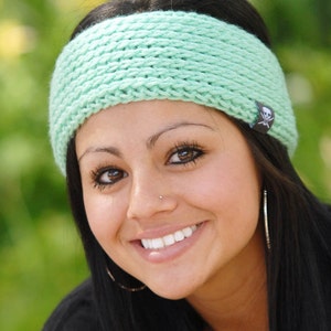 May include: A woman with long black hair is wearing a light green crocheted headband with a skull and crossbones charm. She is smiling and looking at the camera.