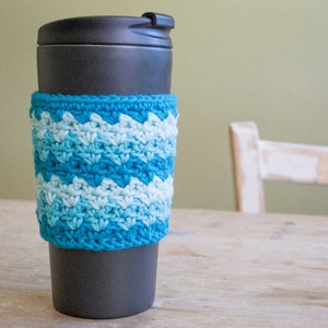 May include: A crocheted coffee cup cozy in a blue and white ombre pattern. The cozy is on a black travel mug.