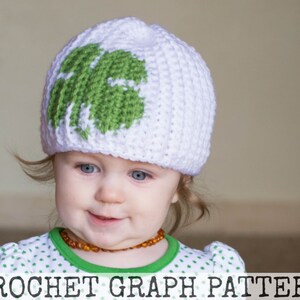 May include: A white crocheted hat with a green four-leaf clover design. The hat is worn by a young child with brown hair and amber beads around their neck.
