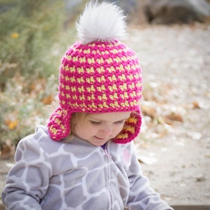 May include: A pink and yellow crocheted hat with ear flaps and a white pom pom. The hat is being worn by a child.
