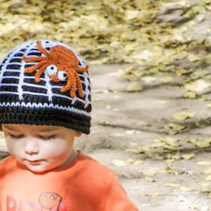 Puede incluir: Un niño lleva un gorro de crochet blanco y negro con forma de telaraña con una araña de crochet marrón y naranja en la parte superior. El gorro tiene un borde negro.