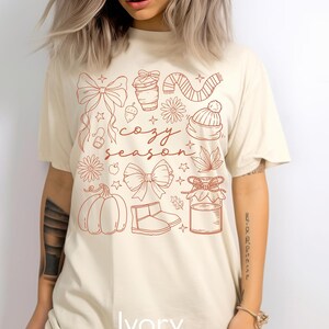 Peut inclure: T-shirt ivoire avec un motif graphique marron repr&eacute;sentant des n&oelig;uds, une tasse de caf&eacute;, une &eacute;charpe, un chapeau, des fleurs, une citrouille et les mots "cozy season". Le mot "Ivory" est imprim&eacute; en bas du t-shirt.