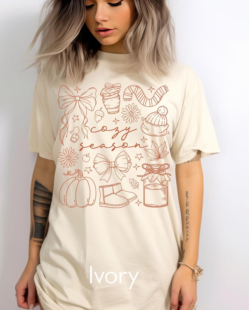 Peut inclure: T-shirt ivoire avec un motif graphique marron repr&eacute;sentant des n&oelig;uds, une tasse de caf&eacute;, une &eacute;charpe, un chapeau, des fleurs, une citrouille et les mots "cozy season". Le mot "Ivory" est imprim&eacute; en bas du t-shirt.