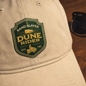 May include: A tan baseball cap with a green and gold patch that reads "Sand Slayer Dune Rider Estd. 1987" and a graphic of a dune buggy. Black sunglasses rest on a wooden surface.