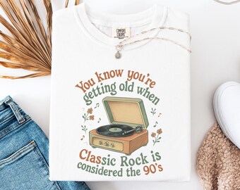 May include: White t-shirt with a retro graphic of a record player and the text "You know you're getting old when Classic Rock is considered the 90's." The design features brown, green, and orange colors.