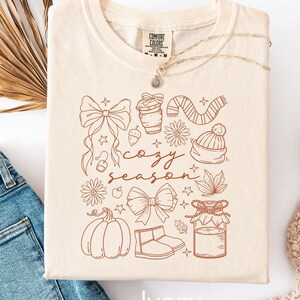 Peut inclure: T-shirt ivoire pli&eacute; avec un motif "Cozy Season" marron, comprenant une citrouille, un n&oelig;ud et une tasse &agrave; caf&eacute;. Le design pr&eacute;sente des illustrations d'automne. Le t-shirt porte une &eacute;tiquette Comfort Colors et le mot "Ivory".
