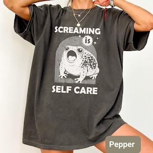 May include: Dark grey t-shirt with the text "SCREAMING IS SELF CARE" and a white graphic of a frog with its mouth open. The shirt is a relaxed fit.