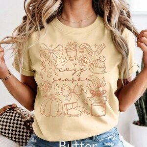 Peut inclure: T-shirt jaune beurre avec un motif rouge d'illustrations sur le th&egrave;me de l'automne, dont une citrouille, un n&oelig;ud, des bottes et les mots "cozy season". Le mot "Butter" est imprim&eacute; en bas.