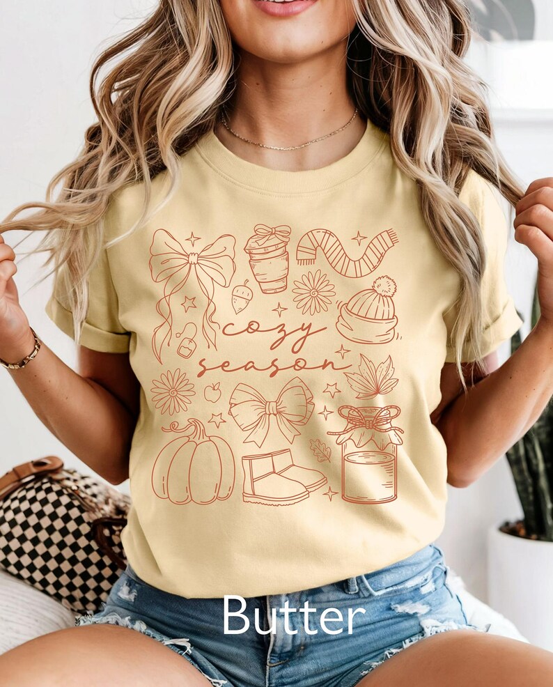 Peut inclure: T-shirt jaune beurre avec un motif rouge d'illustrations sur le th&egrave;me de l'automne, dont une citrouille, un n&oelig;ud, des bottes et les mots "cozy season". Le mot "Butter" est imprim&eacute; en bas.